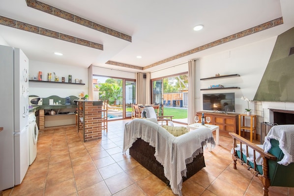 A spacious living area with a sofa, a TV, and a fireplace. There is a kitchen area with a washing machine and a dining area with a table and chairs.