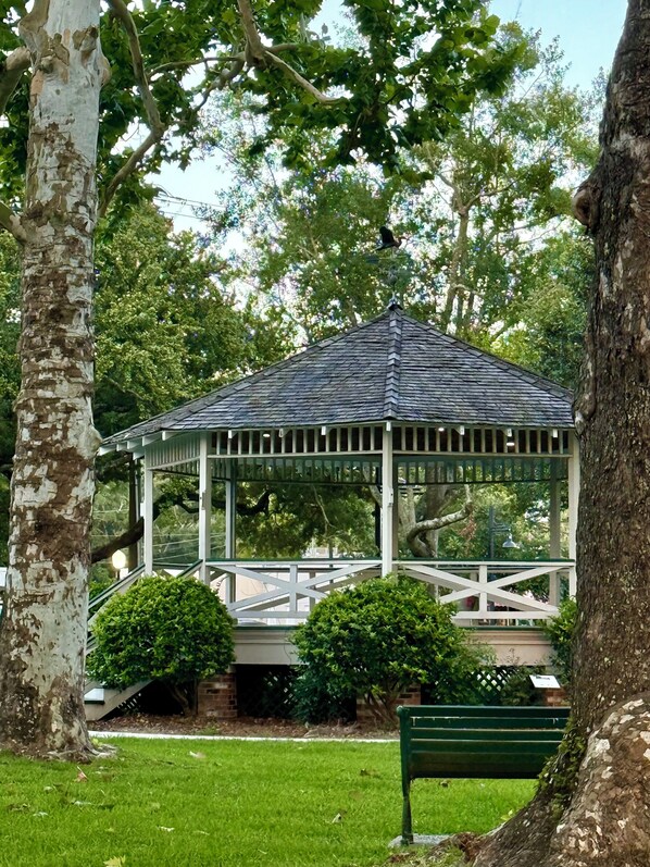 A gazebo with a sloped roof, surrounded by trees and a grassy area.