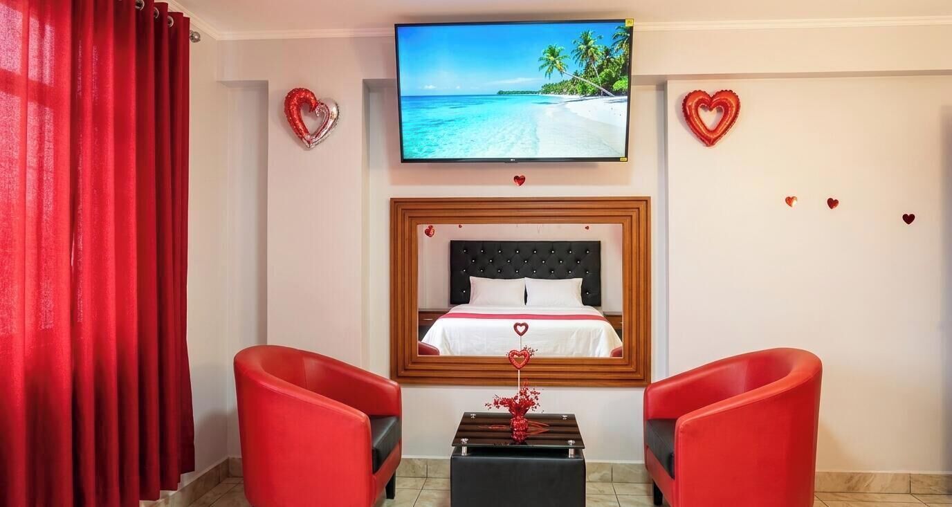 A hotel room with a bed, a TV, red chairs, and decorative hearts on the walls.