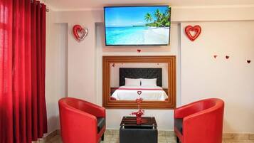A hotel room with a bed, a TV, red chairs, and decorative hearts on the walls.