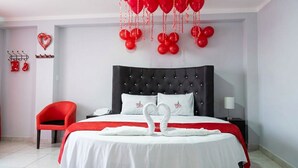 Ein Hotelzimmer mit einem schwarzen, gepolsterten Kopfteil, einem roten Sessel, einem Bett mit weißer Bettwäsche und roten Ballons.