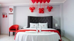 Habitación de hotel con cabecera tapizada negra, una silla roja, una cama con ropa blanca y globos rojos.