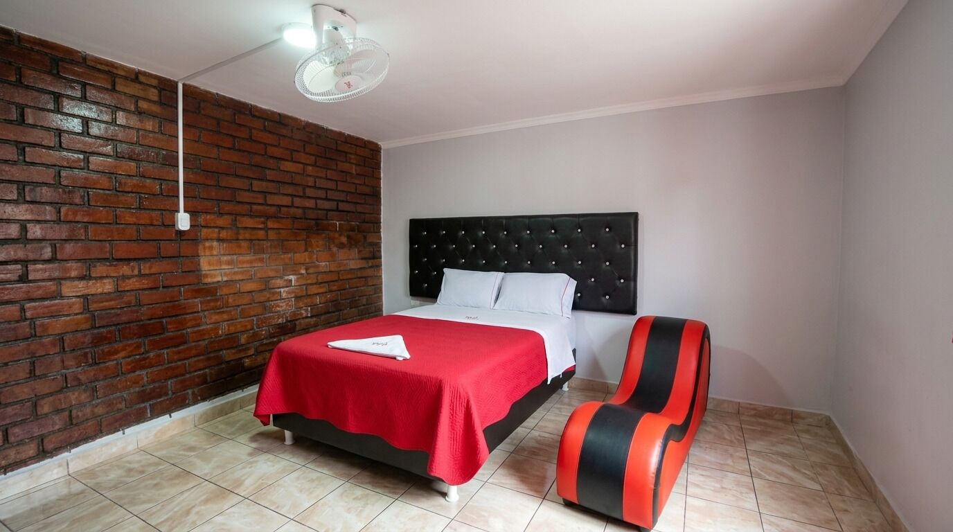 Habitación de hotel con pared de ladrillo, una cama con cubrecama rojo, cabecera negra, una silla roja y negra, y un ventilador de techo.
