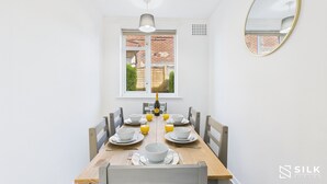 A dining area with a wooden table set for four, white chairs, a window with a view of a brick building, and a wall-mounted mirror. - Modern 3-Bed | Home Near QA Hospital | Sleeps 7 (Cosham)