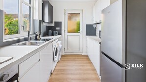 A compact kitchen with a washing machine, a refrigerator, and a window providing a view of greenery outside. - Modern 3-Bed | Home Near QA Hospital | Sleeps 7 (Cosham)