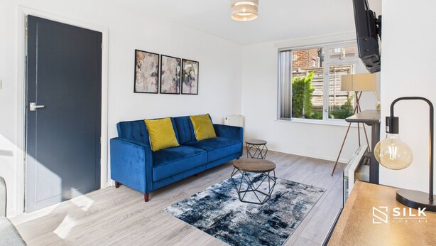 A modern living room with a blue sofa, a coffee table, and a blue and white rug. There is a window with a view of buildings outside. - Modern 3-Bed | Home Near QA Hospital | Sleeps 7 (Cosham)