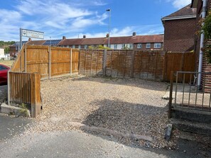 A closed area with a wooden fence, gravel ground, and residential buildings in the background. - Modern 3-Bed | Home Near QA Hospital | Sleeps 7 (Cosham)