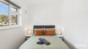 A neatly made bed with a green bedspread, two pillows, and a rolled towel. There is a window with a view of houses outside. - Modern 3-Bed | Home Near QA Hospital | Sleeps 7 (Cosham)