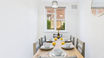 A dining area with a wooden table set for four, white chairs, a window with a view of a brick building, and a wall-mounted mirror.