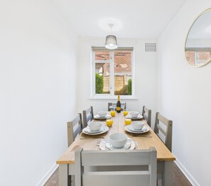 Un coin repas avec une table en bois dressée pour quatre personnes, des chaises blanches, une fenêtre donnant sur un bâtiment en briques et un miroir fixé au mur.