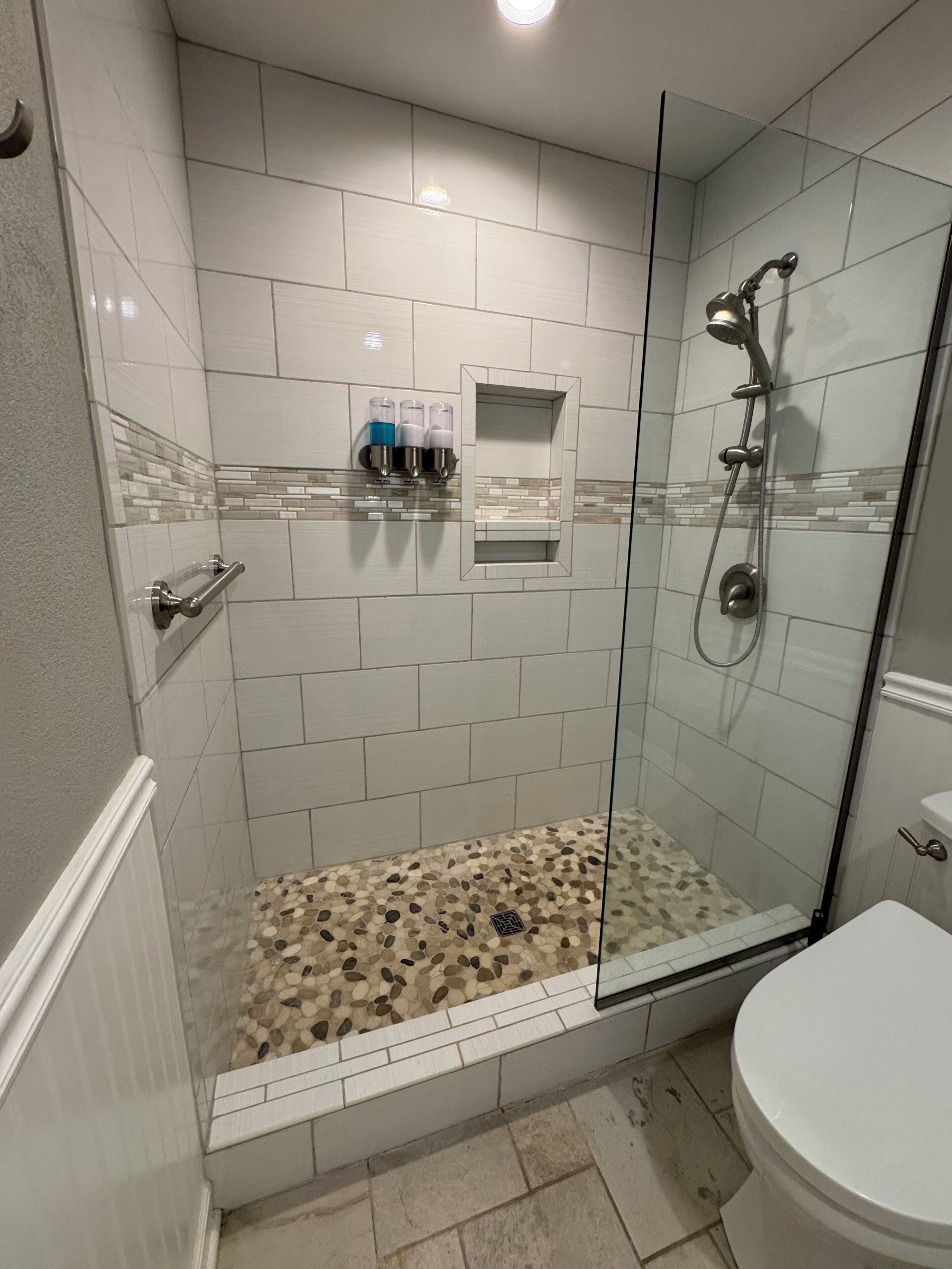 A shower area with a glass door, white tiled walls, a small shelf with toiletries, and a pebble-patterned floor.