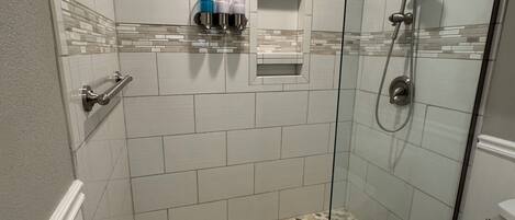 A shower area with a glass door, white tiled walls, a small shelf with toiletries, and a pebble-patterned floor.