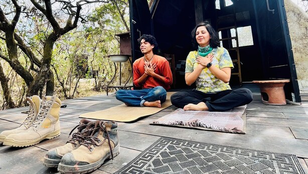 Two people meditating on outdoor mats with a rustic shelter and trees in the background.