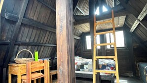 A rustic attic room with wooden beams, a loft bed, a wooden ladder, and a table with a basket and a green water bottle.