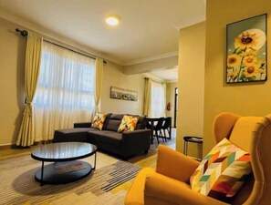 Ein Wohnzimmer mit einer Couch, einem runden Couchtisch und einem gelben Sessel. An der Wand hängt ein Gemälde mit Sonnenblumen.