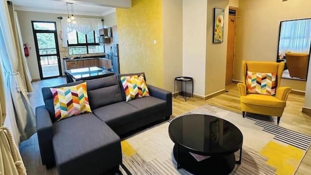 A modern living room with a sofa, armchairs, and a coffee table. There is a kitchen in the background with a dining table and chairs.