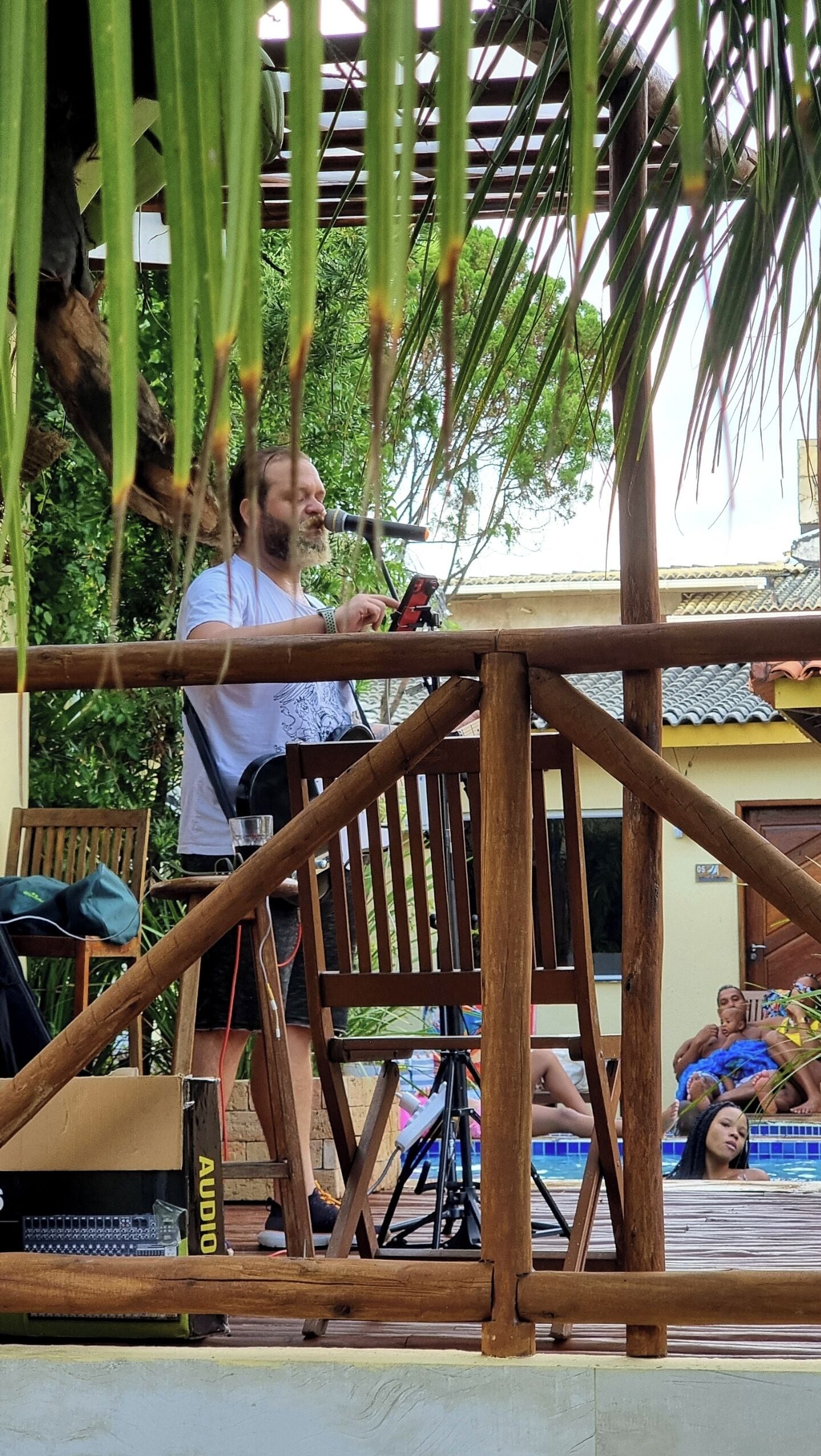 Una persona de pie sobre una terraza de madera, con un micrófono, una silla y una mesa en primer plano, y una piscina con gente en el fondo.