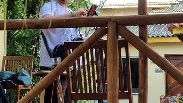 A person standing on a wooden deck with a microphone, a chair, and a table in the foreground, and a pool with people in the background.