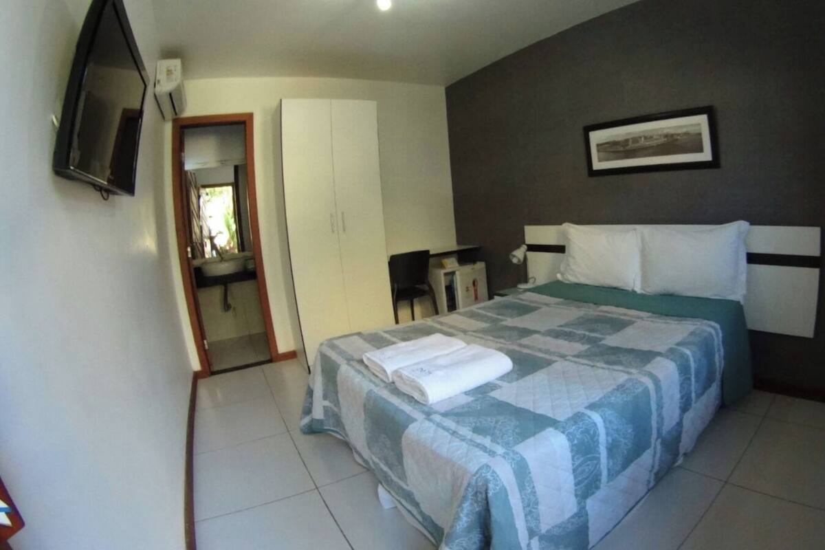Une chambre à coucher comprenant un lit, un bureau, une chaise, un téléviseur et une salle de bain visible à travers une porte ouverte.