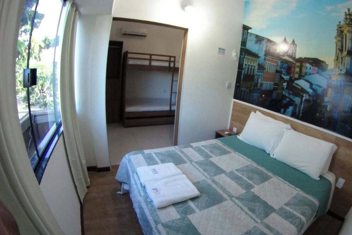 Une chambre d’hôtel avec un lit, une fenêtre avec des rideaux, un tableau représentant un paysage urbain accroché au mur et une porte donnant sur une autre pièce.