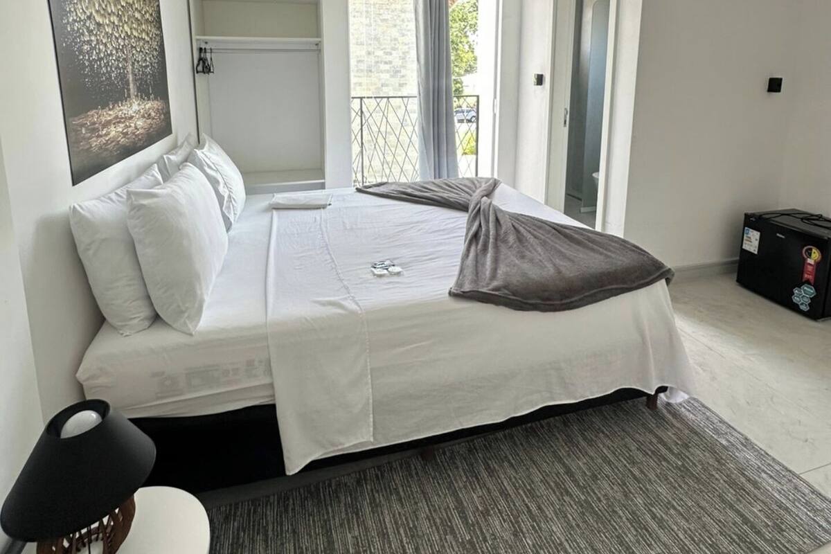 Une chambre moderne avec un grand lit, une table de chevet, une chaise, une fenêtre avec des rideaux et une vue sur l’extérieur.