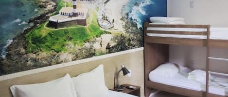 Hotelkamer met een bed, een stapelbed, nachtkastje, lamp en een muurschildering van een kuststadsgezicht.