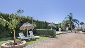 Een geplaveid pad met ligstoelen en rieten parasols in een aangelegde tuin.