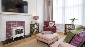 A living room with a fireplace, a television mounted above it, a red armchair, a pink ottoman, and a large window with curtains. - Scottish Thistle - 1 bedroom with optional sofa bed (Inverness)