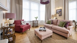 A well-lit living room with a red armchair, a pink sofa, and a matching ottoman. There is a wooden side table with a lamp and a tray of books. - Scottish Thistle - 1 bedroom with optional sofa bed (Inverness)