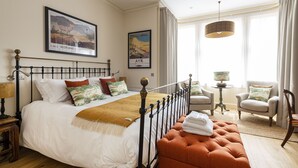Une chambre d’hôtel bien éclairée, dotée d’un grand lit, d’un coin salon avec un fauteuil et une table, et de deux tableaux encadrés accrochés au mur.