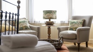 Une pièce confortable avec un lit, une chaise, une table avec une lampe et une fenêtre ornée de rideaux légers.