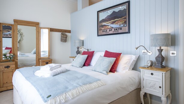 En snyggt bäddad säng med vita och blå sängkläder, en inramad bild av ön Skye och en sänglampa på ett litet bord bredvid sängen.