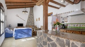 A room with a stone counter, wooden beams, a stone fireplace, a bed with a blue cover, a television, and a kitchen area with a clock. - Casa Marilo country house with pool in Conil de la Frontera (Conil de la Frontera)