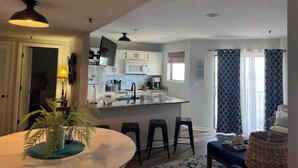 Eine moderne Küche mit weißen Schränken, einer zentralen Kochinsel und Barhocker. Es gibt einen Essbereich mit rundem Tisch und Stühlen, sowie eine Wohnbereich mit einer Couch und einem großen Fenster.