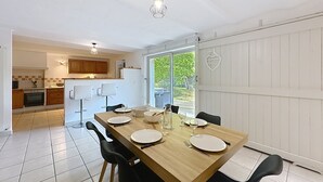 Un coin repas avec une table en bois dressée pour quatre personnes, des chaises noires et, au fond, une cuisine dotée de meubles en bois et d’un réfrigérateur blanc.