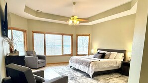 Quarto com cama, duas poltronas, ventilador de teto e janelas amplas com persianas.