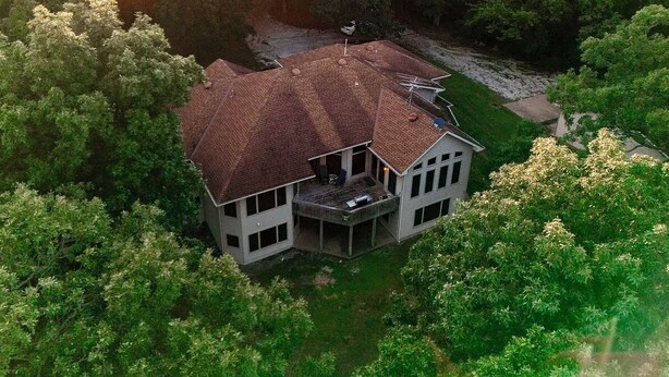 Uma casa de dois andares com uma varanda grande e uma área externa coberta, cercada por árvores densas.
