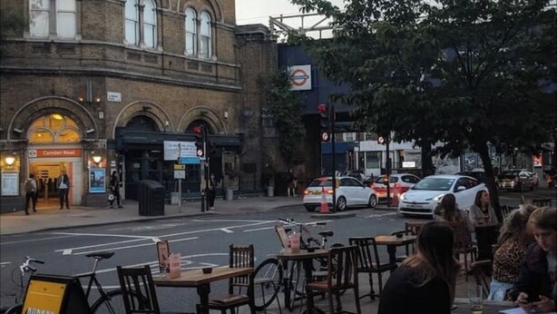A street view with outdoor seating at a restaurant, a tram stop, and a building with a sign for 'Camden Rail'.
