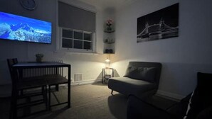 A dimly lit room with a bed, a TV mounted on the wall, a window, and a small table with a lamp.