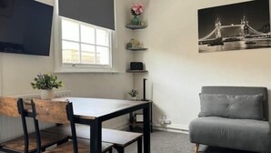 A room with a dining table, chairs, a grey sofa, a TV mounted on the wall, a window, and a picture of a bridge.