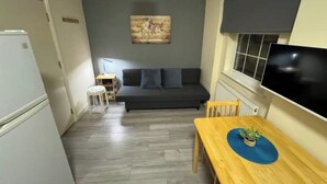 A compact living space with a sofa, a small table with a plant, a TV mounted on the wall, and a refrigerator.
