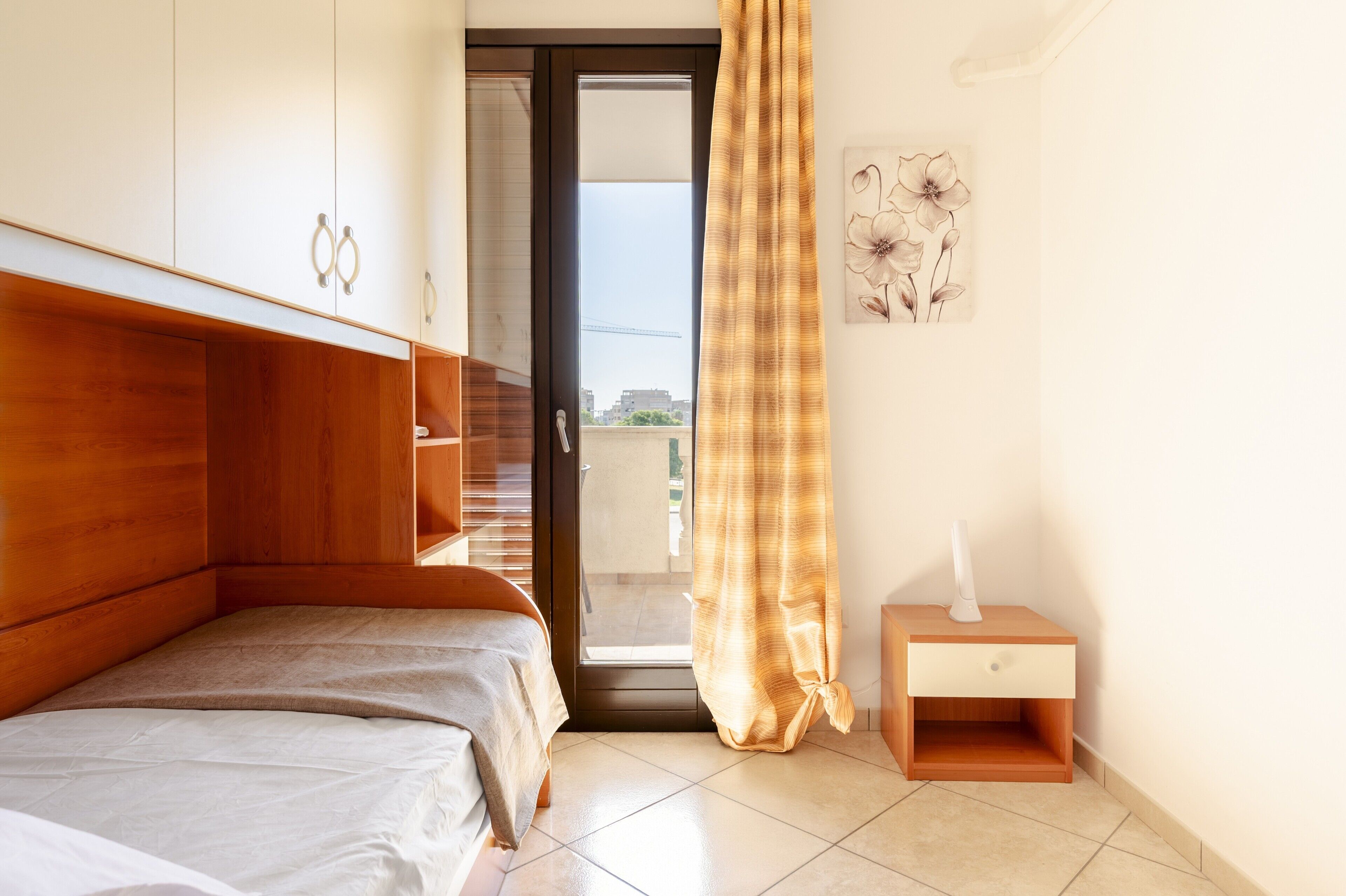 A bedroom with a bed, a nightstand, a door leading to an outdoor balcony, and a wall with a floral painting.