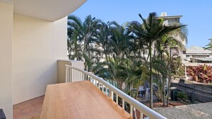 Uma varanda com mesa de madeira e grade branca, com vista para um pátio com palmeiras e um edifício ao fundo.