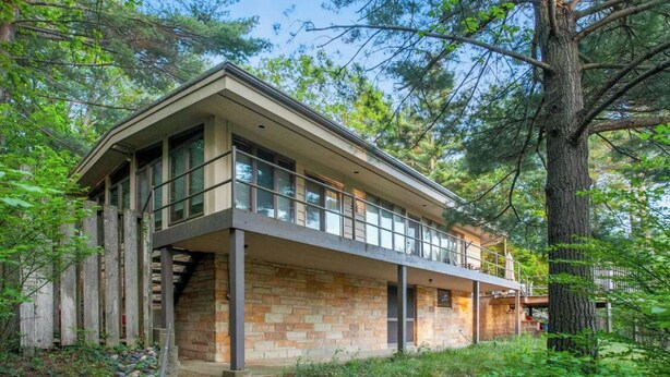 A two-story house with a stone facade, large windows, and a balcony overlooking a forested area.