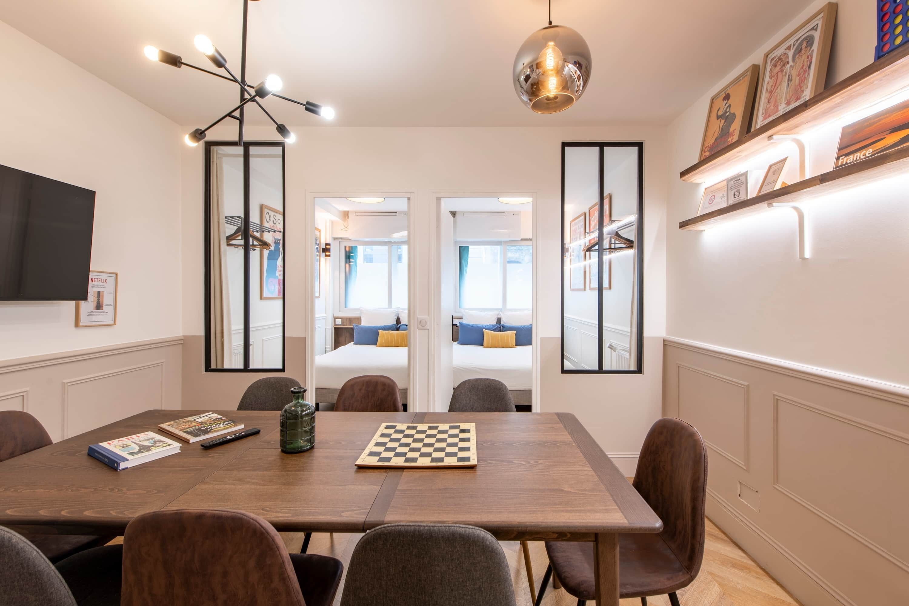 A modern hotel room with a wooden dining table, chairs, a chessboard, and a large mirror.