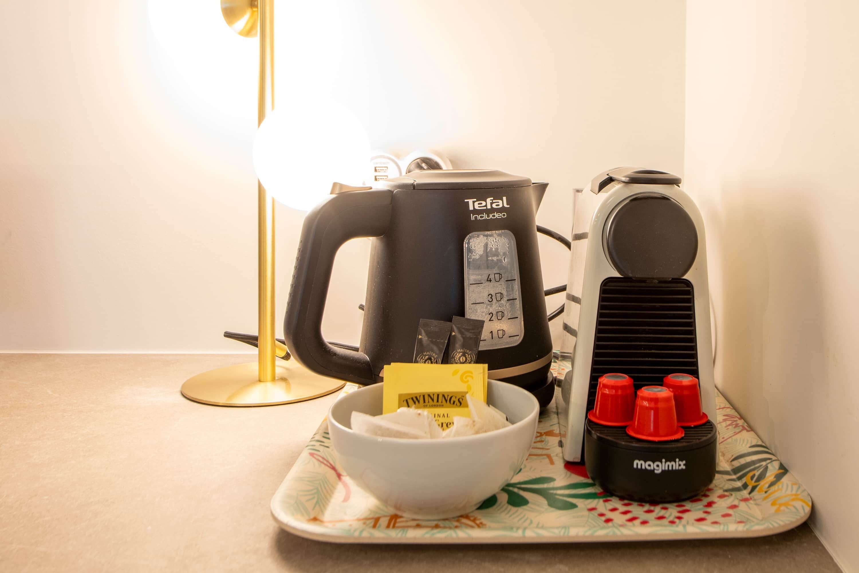 A kitchen area with a coffee maker, a kettle, a bowl with tea bags, and a tray with red cups.