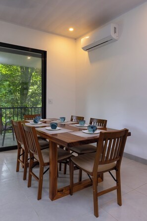 A dining area with a wooden table and chairs, a glass window with a view of trees, and an air conditioning unit on the wall. - Villa del Río (Savegre)