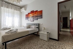 A bedroom with a bed, a nightstand, a wall-mounted air conditioner, and a painting of a sunset.