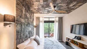 Une chambre avec un mur en pierre, un ventilateur de plafond, un lit avec du linge de lit blanc, un téléviseur fixé au mur et un balcon offrant une vue sur la verdure.