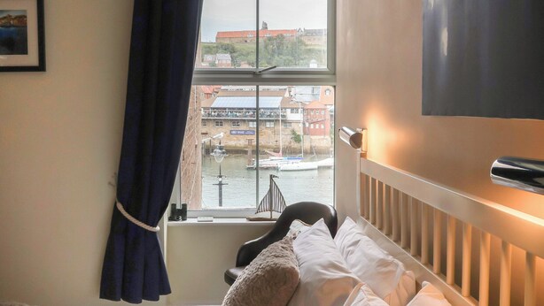 A hotel room with a bed, a window with a view of a canal and buildings, a chair, and a wall-mounted light fixture. - Jolly Roger (Whitby)